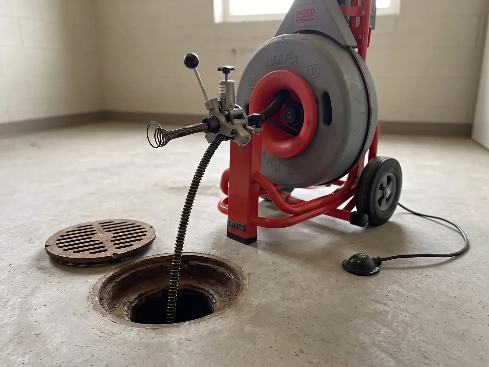 A red cleaning machine on a bare concrete floor with its cable running down an open floor drain, and the drain grate lying beside it